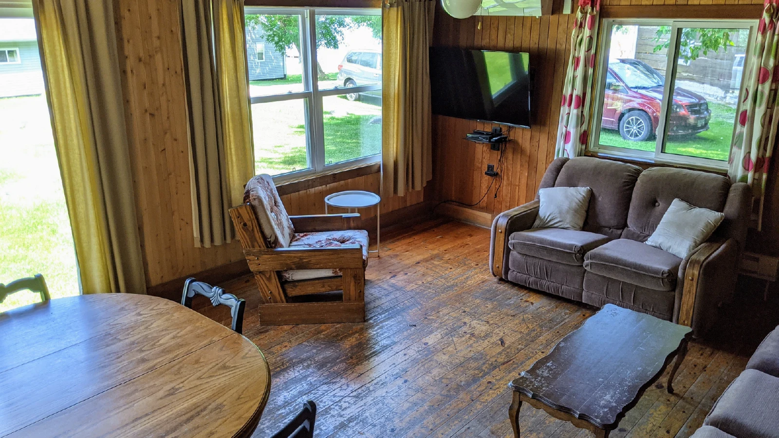 Pigeon Lake Resort Cottage Six.&nbsp; Open-concept cottage living and dining area with wood interior, sofa, armchair, and large windows.